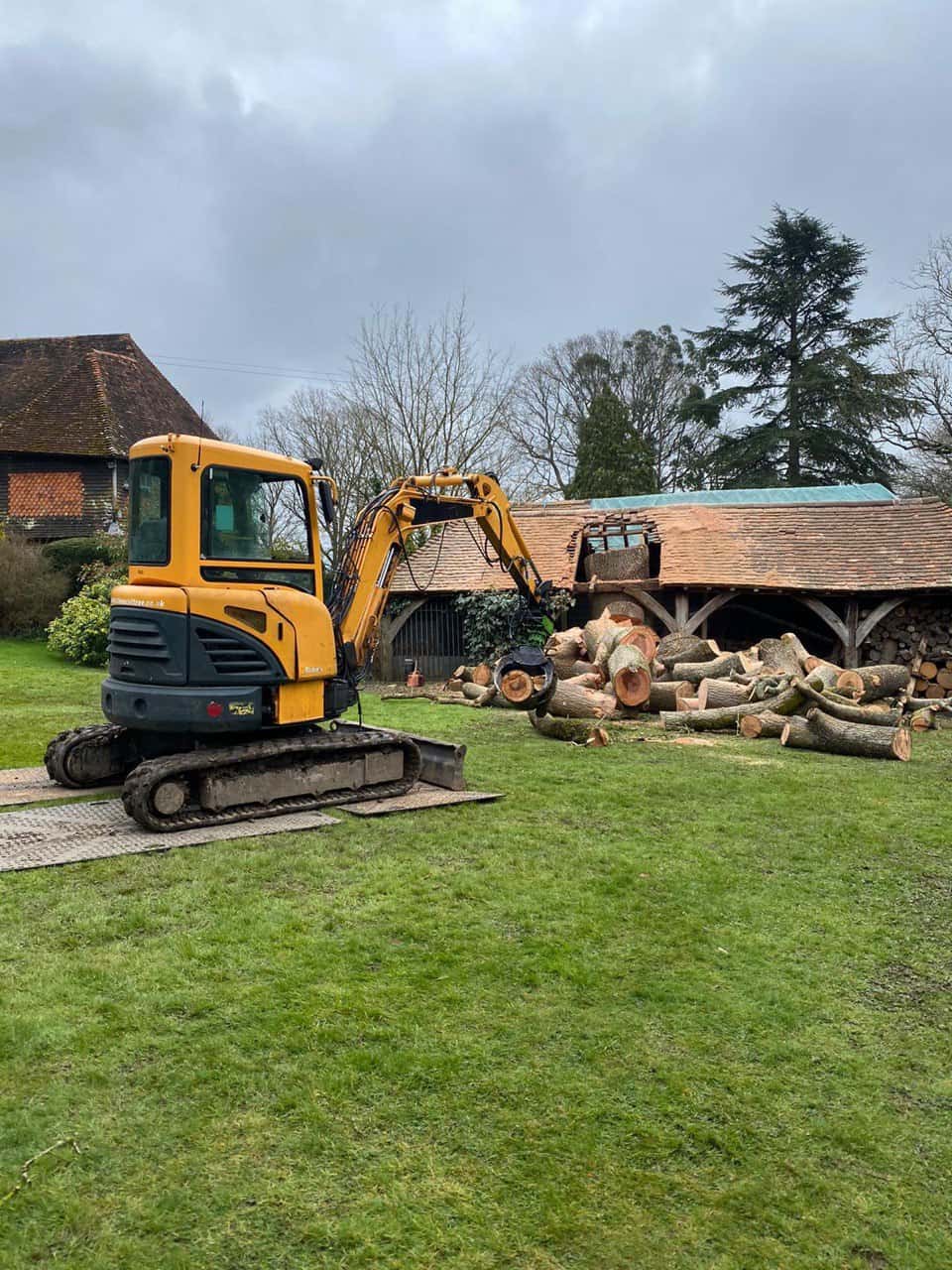 This is a photo of a tree which has grown through the roof of a barn that is being cut down and removed. There is a digger that is removing sections of the tree as well. Eastwood Tree Surgeons